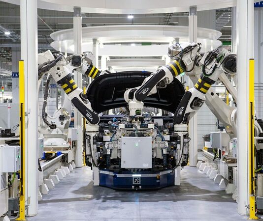 Dördüncü Sanayi Devrimi olarak da anılan günümüz endüstriyel dönüşümünde robotik teknolojiler, iş dünyasının dinamiklerini radikal bir biçimde yeniden şekillendiriyor. 18. yüzyılda buhar gücünün öncülük ettiği ilk sanayi devriminden bu yana, üretim teknikleri sürekli olarak evrildi. Ancak hiçbir dönem, günümüzdeki kadar hızlı ve kapsamlı bir değişim yaşamadı. Robotlar, artık sadece otomotiv fabrikalarında değil, sağlık, tarım, lojistik ve daha birçok sektörde üretkenliğin temel direği haline geldi. Bu gelişmeler, robotik sistemlerin yalnızca iş süreçlerini değil, aynı zamanda iş gücü yapısını, ekonomik modelleri ve toplumsal yapıyı da etkilediğini ortaya koyuyor. Konulu bir haber görseli.